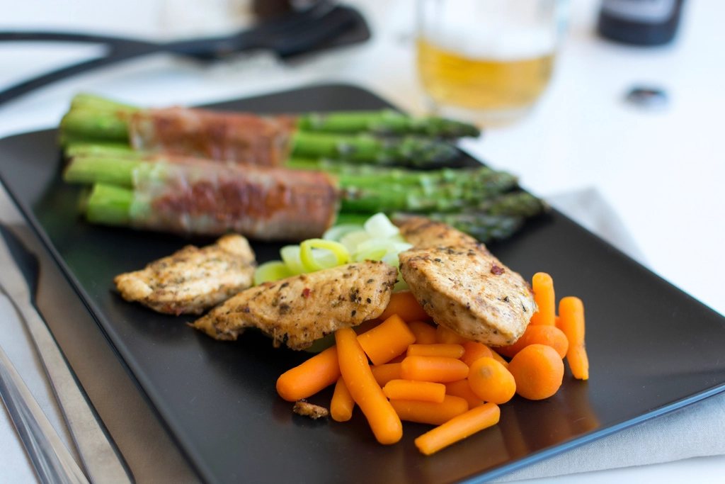 A plated meal of seasoned chicken breast, bacon-wrapped asparagus and baby carrots on a dark tray
