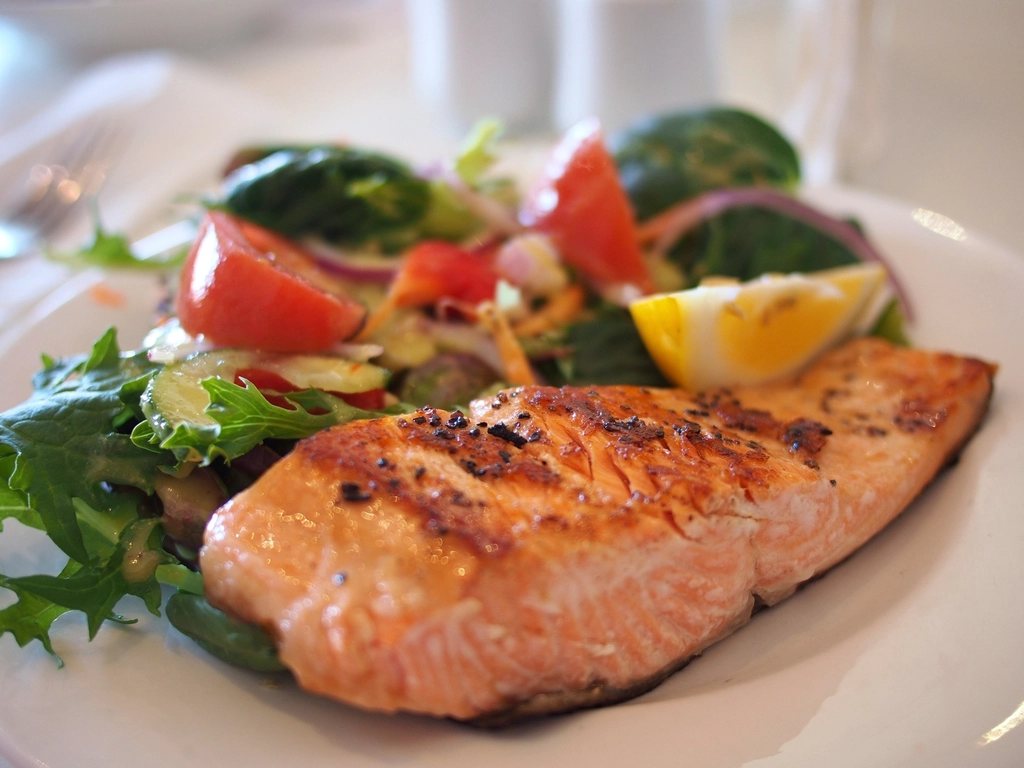 A grilled salmon fillet on a white plate with a mixed salad of tomato, cucumber, red onion and a lemon wedge