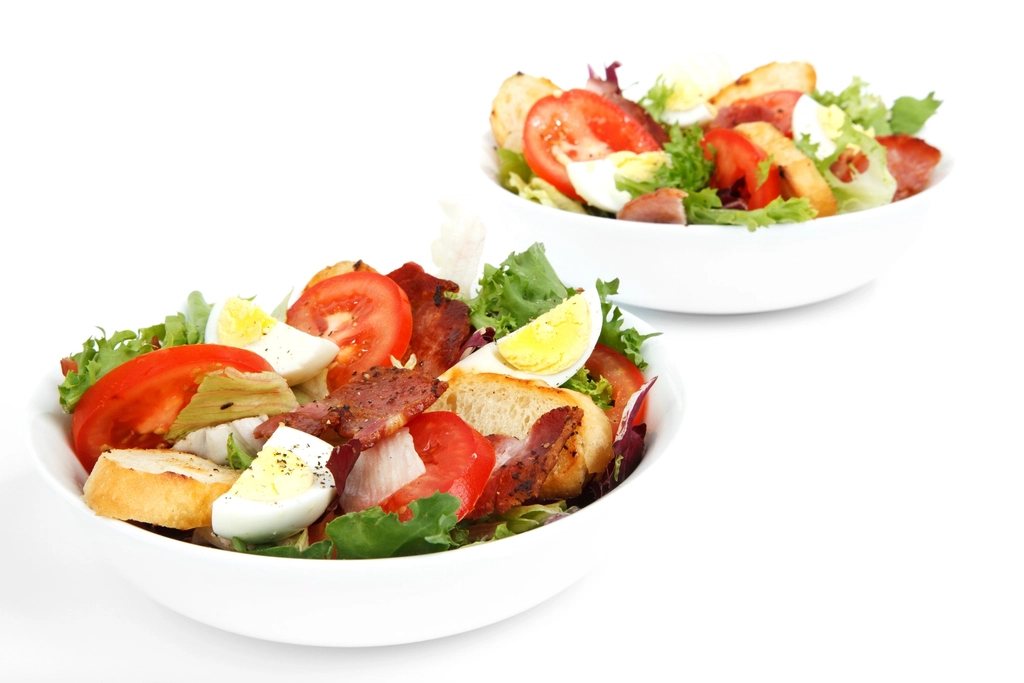 Two white bowls of salad with tomato, boiled egg, bacon, lettuce and croutons